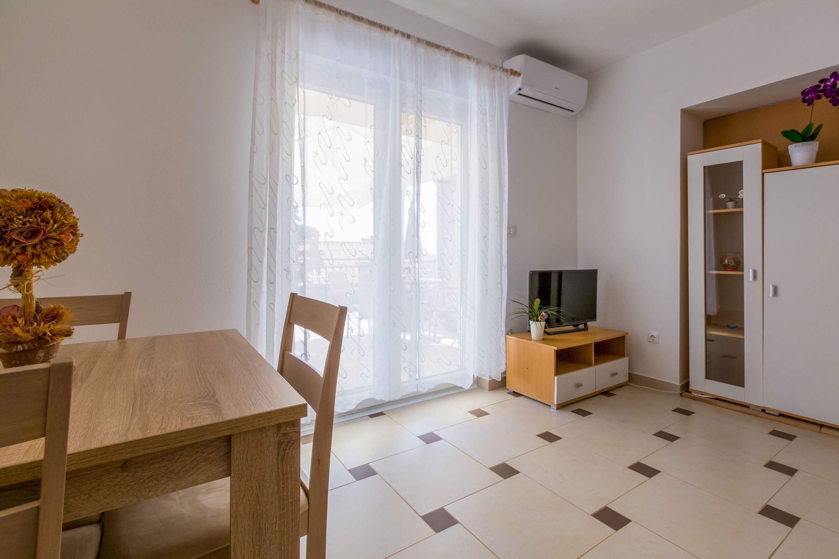 A room with a wooden dining table, chairs, a TV on a wooden stand, a cabinet with shelves, and a large window with sheer curtains.