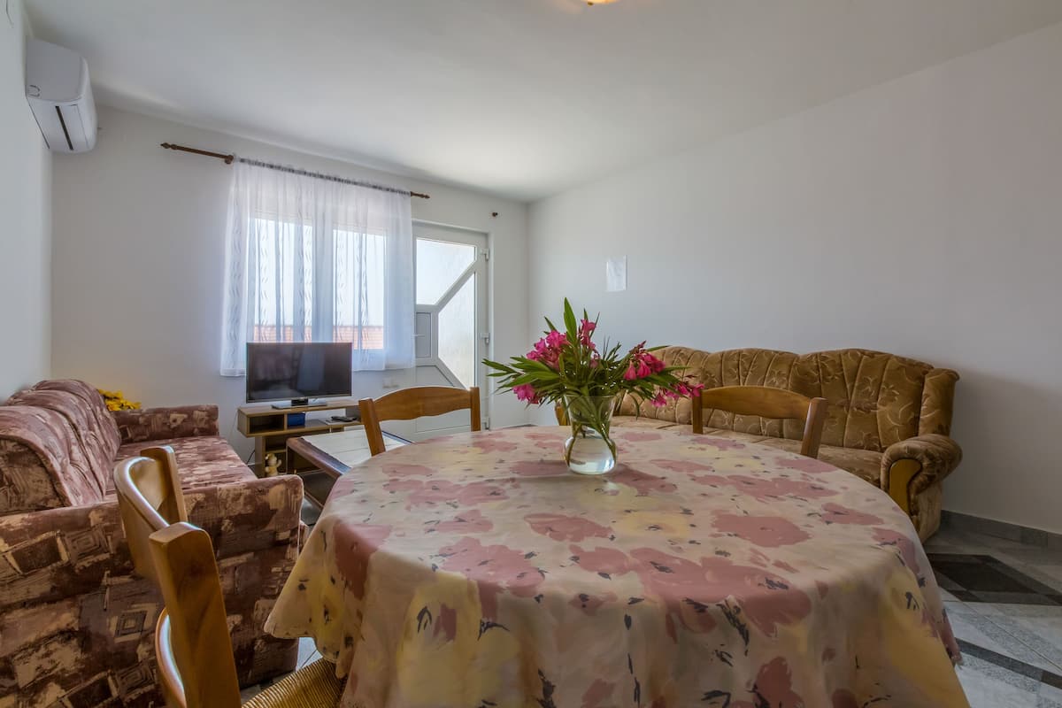 A living room with a round table, floral tablecloth, and a vase of flowers. There is a sofa and a television in the background.