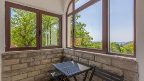 Ein Eckzimmer mit Tisch und Stühlen, große Fenster mit braunen Rahmen und Blick auf Grünflächen und Hügel.