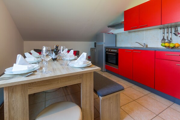 A dining area with a wooden table set for four, a red kitchen with modern appliances, and a tiled backsplash. - Apartment Linda (Grizane)