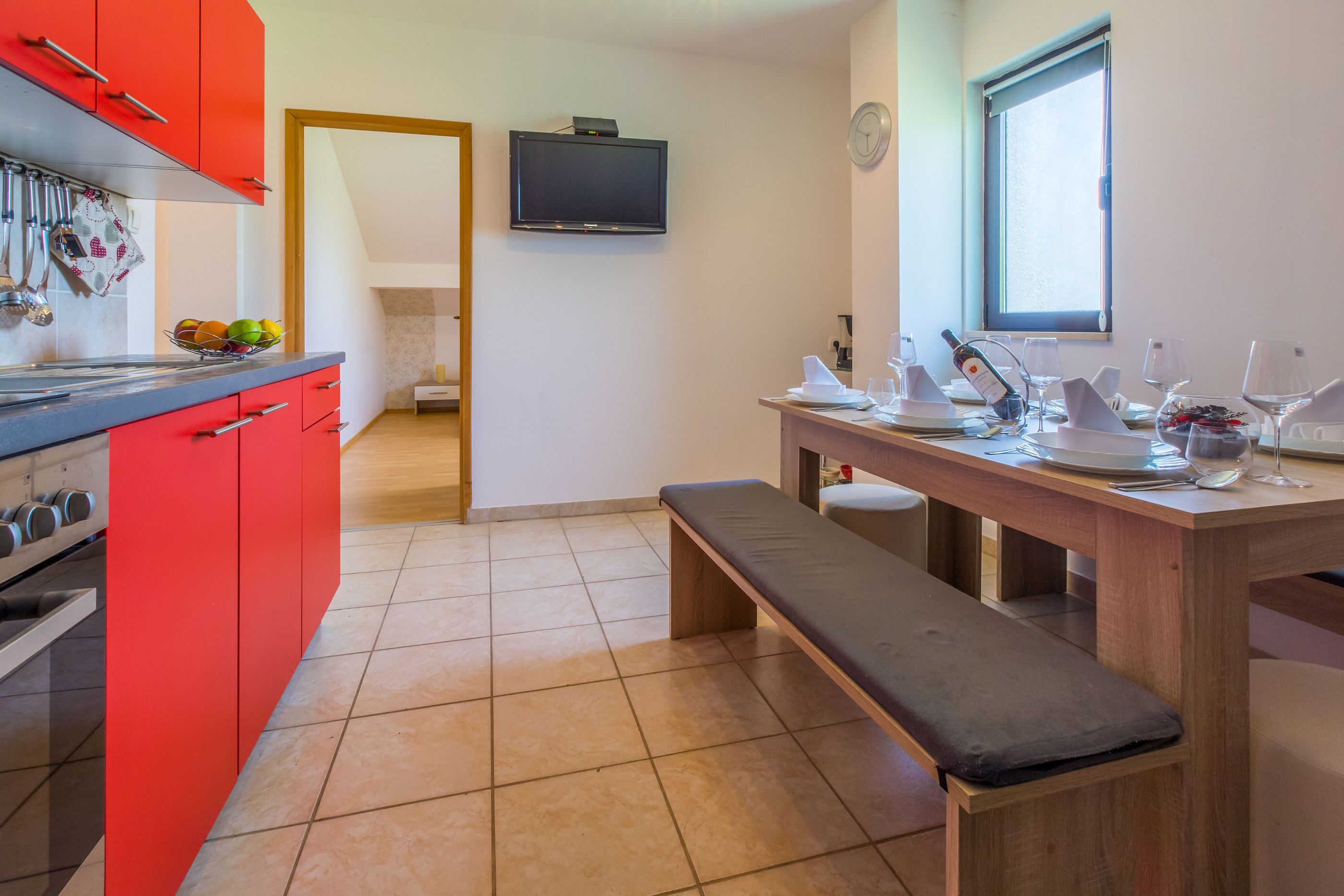 A modern kitchen with red cabinets, a wooden dining table set for two, and a TV mounted on the wall.