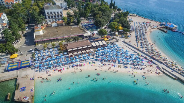 An aerial view of a beach with numerous umbrellas and people. There are buildings and a hotel in the background. - Apartments Marija (Selce)