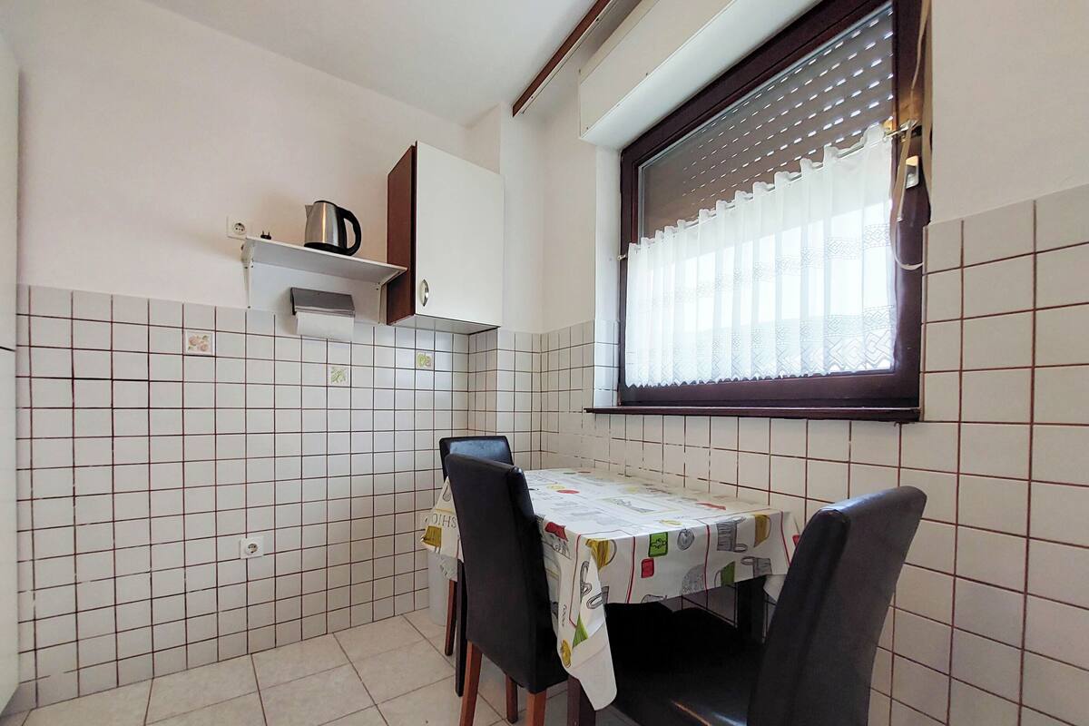 Een kleine keuken met een betegelde spatwand, een raam met zonwering, een tafel met tafelkleed en twee stoelen.
