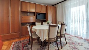 A dining area with a round table, chairs, a television, wooden cabinets, and a patterned rug. - Apartments Marija (Selce)