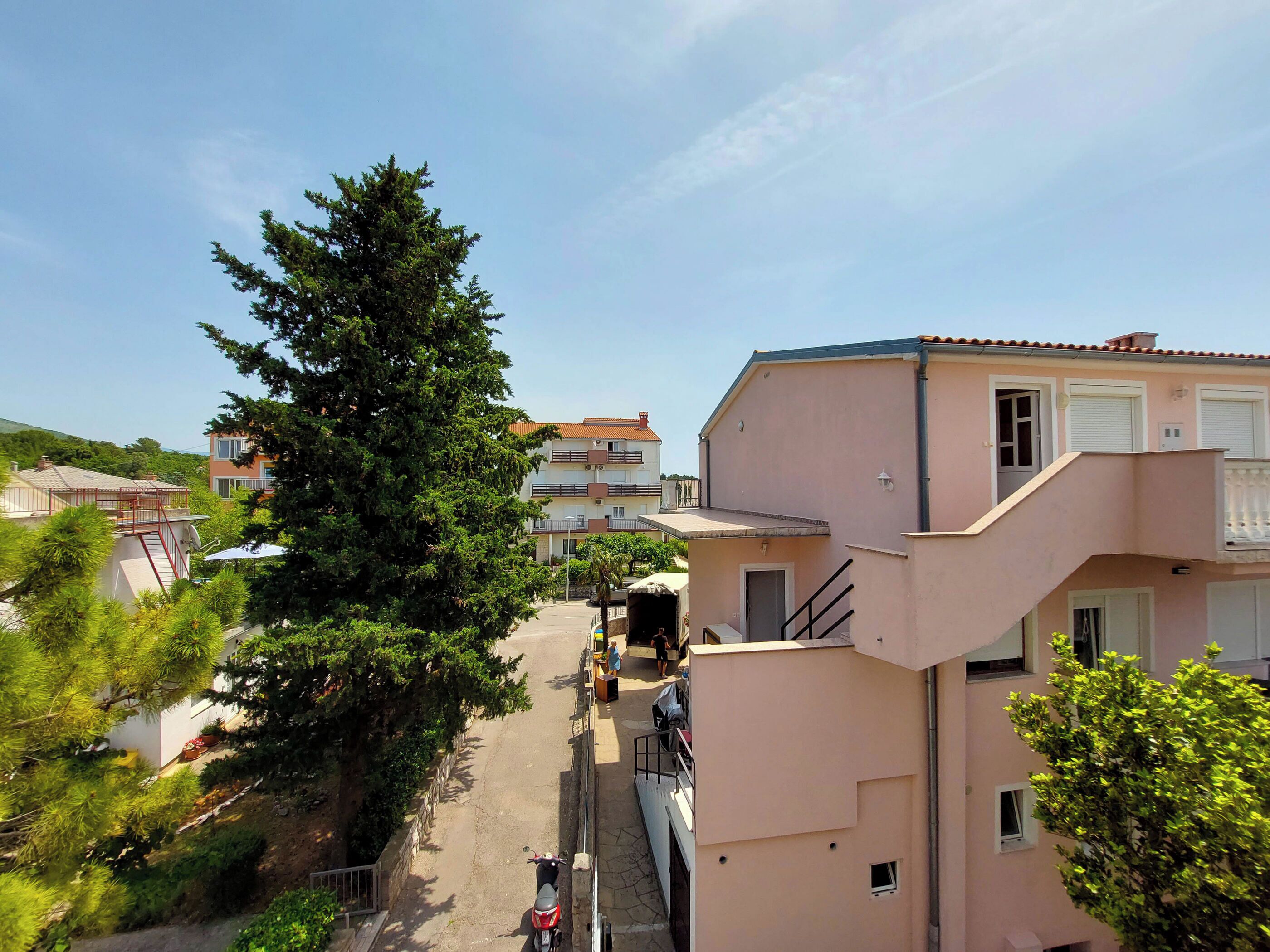 A residential area with a narrow street, multi-story buildings, and a large tree.