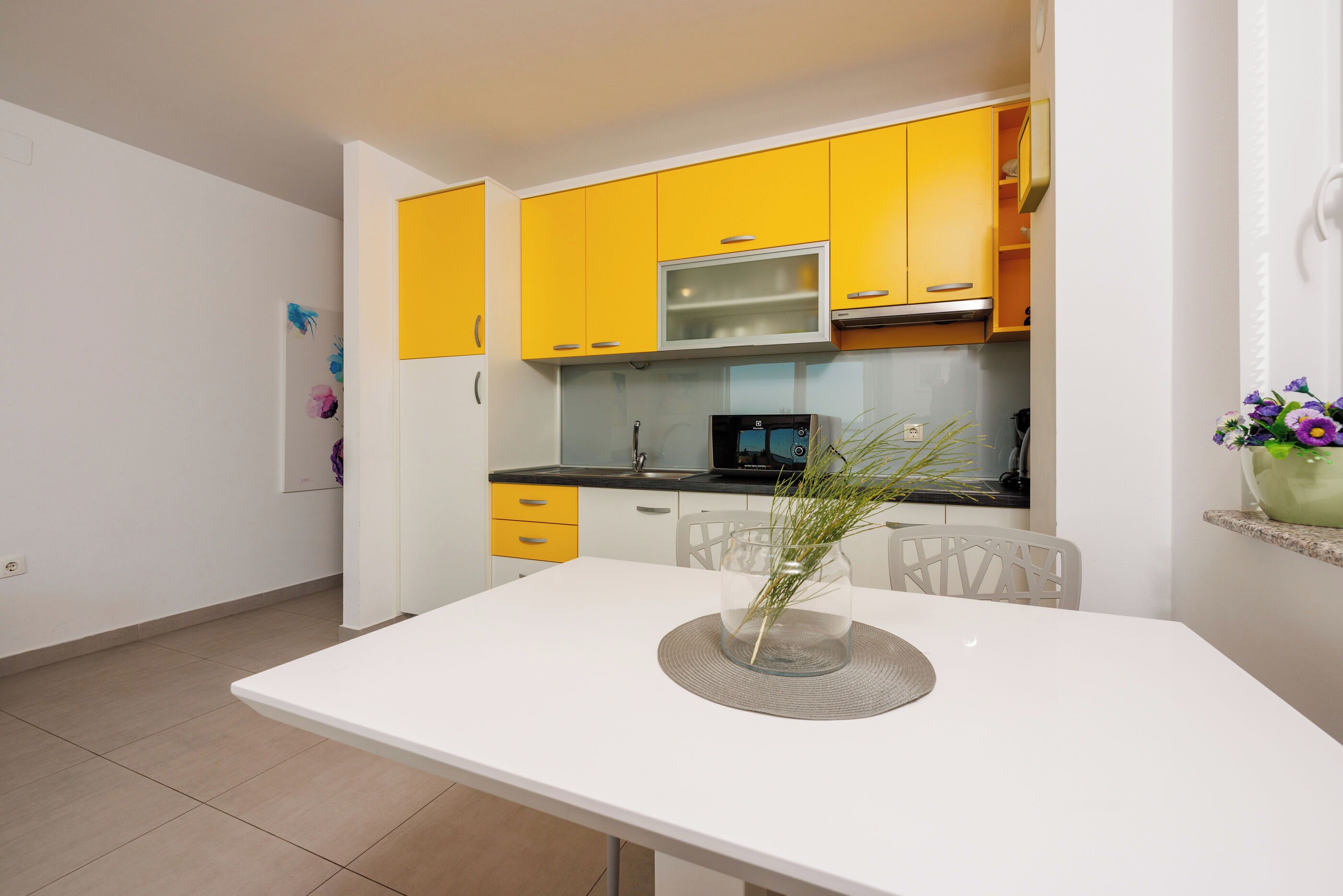 A modern kitchen with yellow cabinets, a white countertop, and a dining table with a vase of flowers.