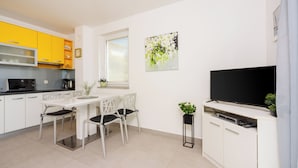 Een moderne keuken met witte kastjes, een eettafel met stoelen, een zwarte televisie op een witte kast en een raam met zonwering.