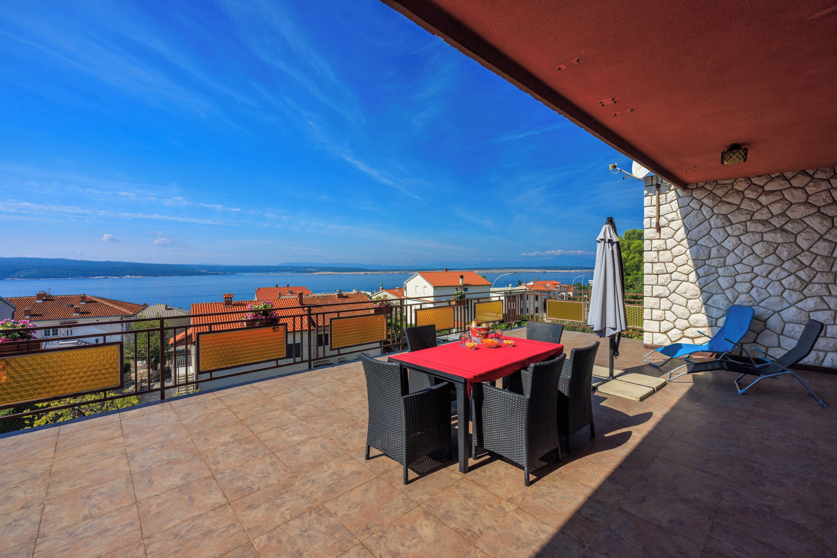 Eine Terrasse mit einem roten Tisch und Stühlen, einer Steinsäule und Blick auf das Meer.