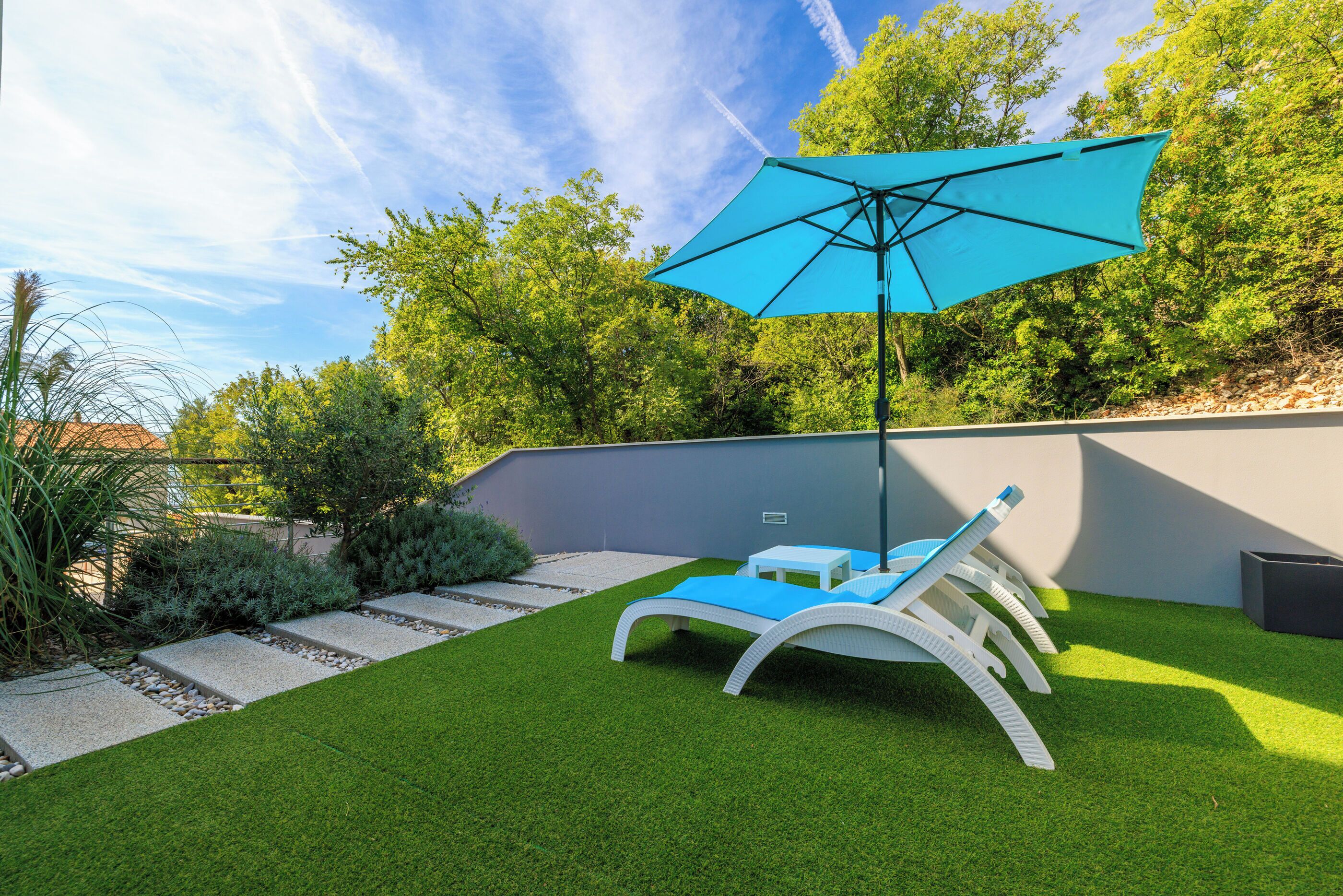A backyard with artificial grass, a white lounge chair, a blue umbrella, and a small table.