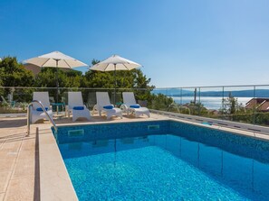 A swimming pool with lounge chairs and umbrellas, surrounded by a glass railing, overlooking a body of water and distant landscape.