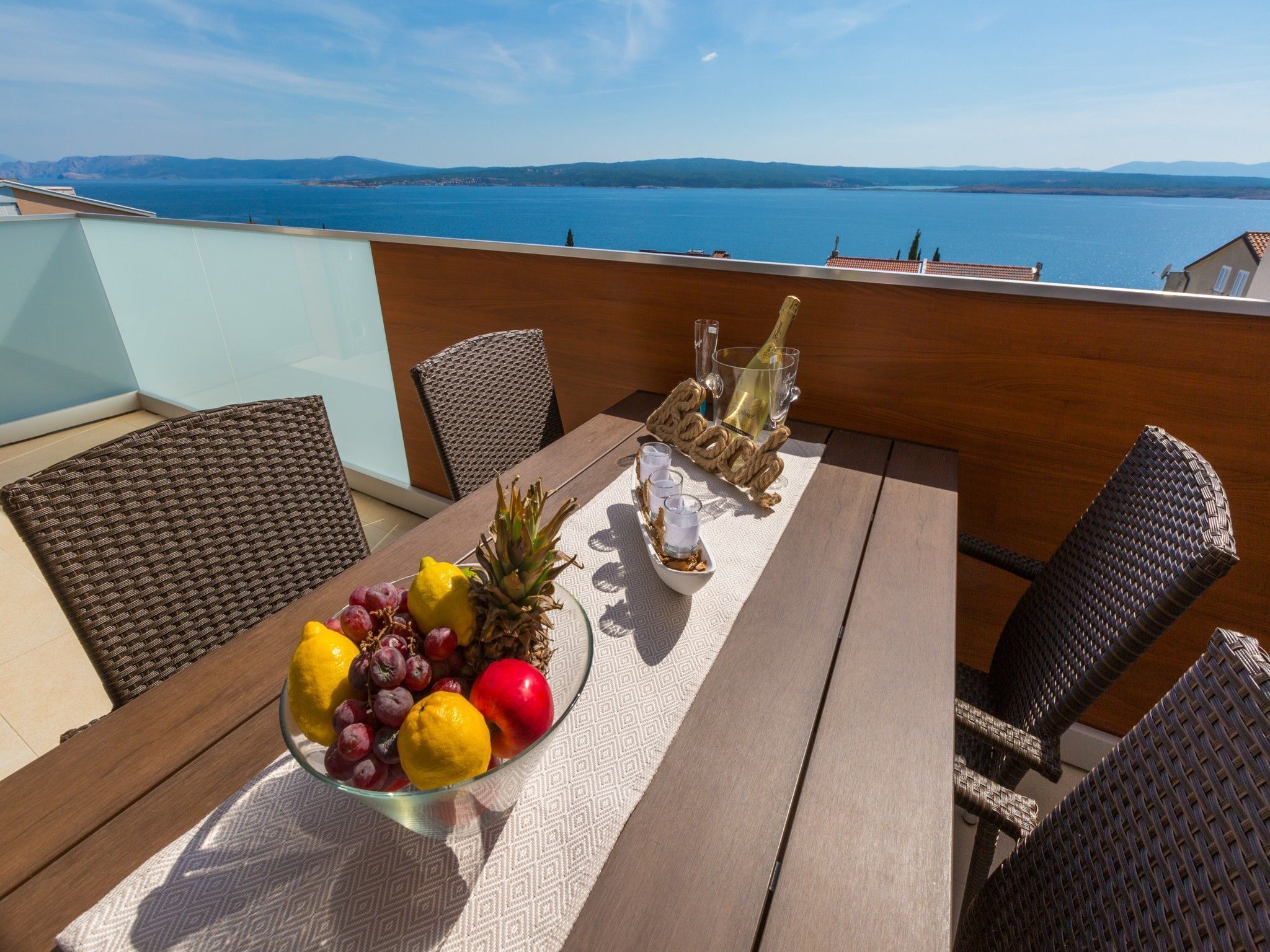 A balcony with a table set for two, featuring a fruit bowl, a bottle of champagne, and a view of the sea.