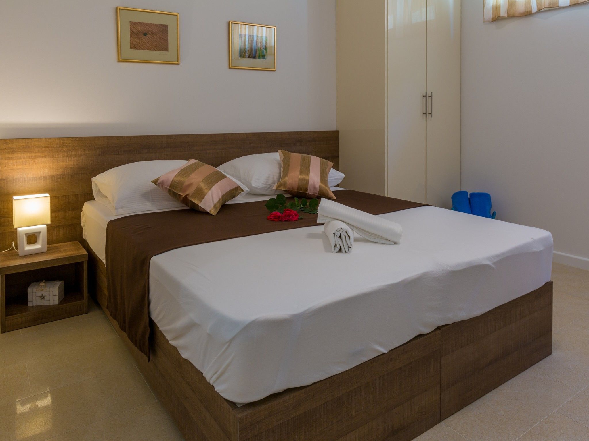 A neatly made bed with white linens, a wooden headboard, and two pillows. There is a nightstand with a lamp and a small box.