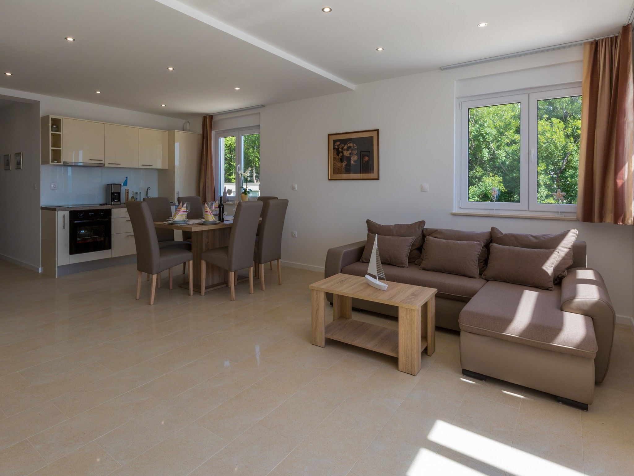 A modern living room with a sofa, a coffee table, and a dining area with chairs and a table. There is a kitchen in the background with cabinets and a stove.
