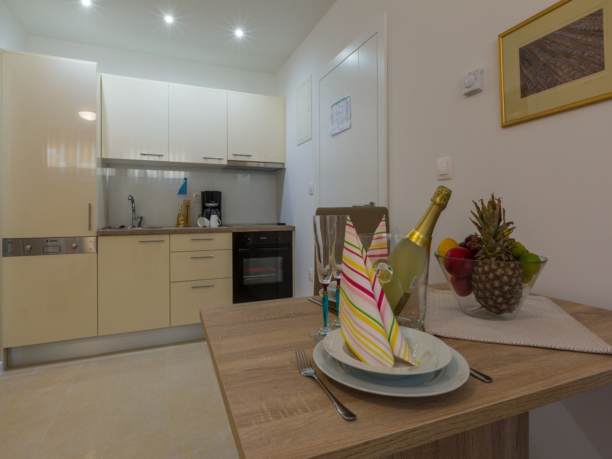 A modern kitchen with white cabinets, a built-in oven, and a coffee maker. There is a dining table with a bottle of champagne, a pineapple, and a bowl of fruit.