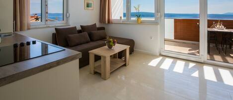 Un salon moderne comprenant un canapé, une table basse et un coin cuisine avec une cuisinière. De grandes fenêtres offrent une vue sur la mer, et un balcon abrite une table à manger.