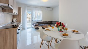 Una cocina moderna con armarios de madera, un comedor con mesa y sillas blancas, y una sala de estar con sofá y vistas a través de un ventanal.