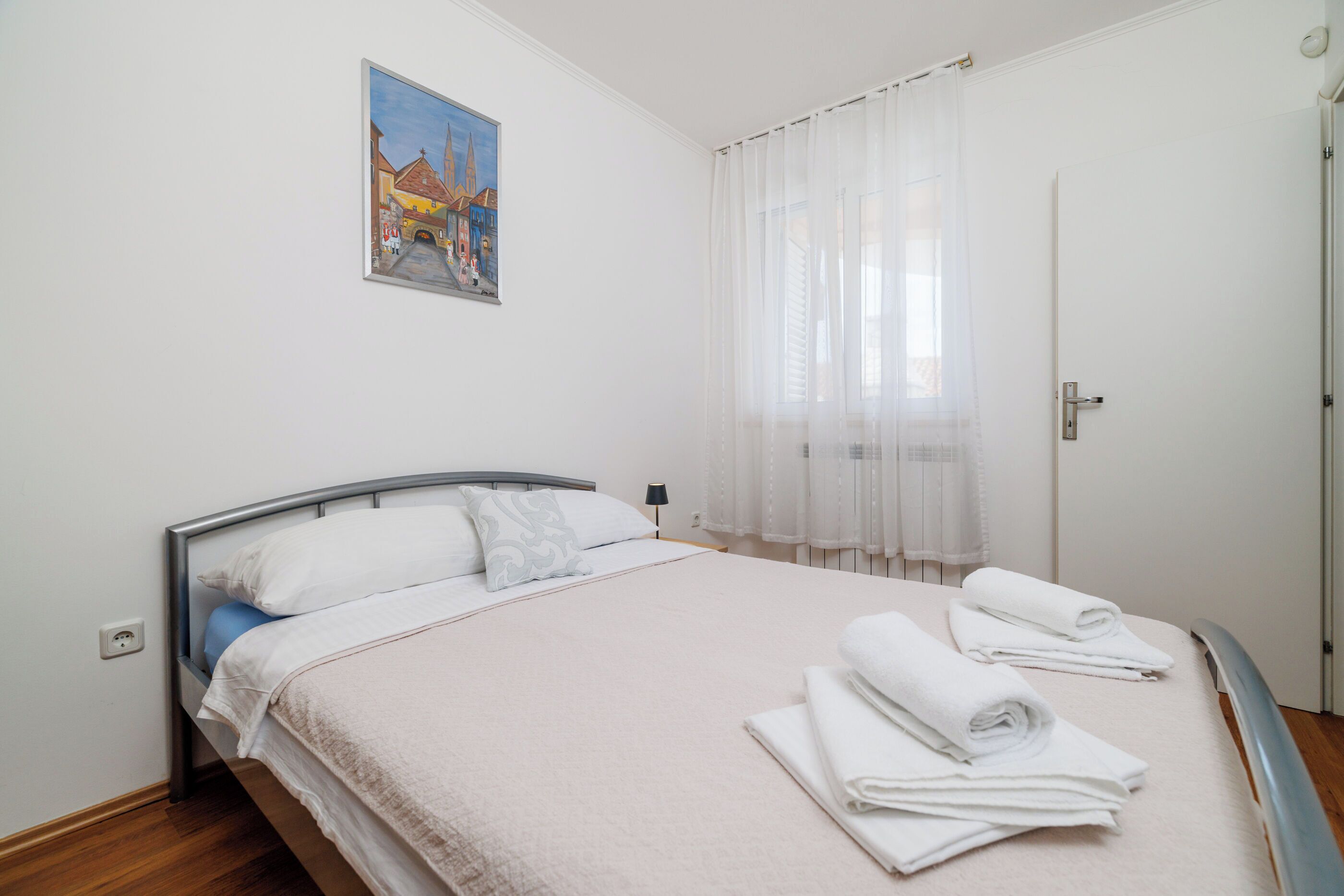 A bedroom with a bed, a painting on the wall, a door, and a window with curtains.