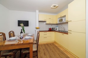 A dining area with a wooden table set for two, a television mounted on the wall, and a kitchen with yellow cabinets and a microwave. - Apartments Carmen (Crikvenica)