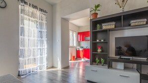 A modern living room with a grey sofa, a flat-screen TV on a black stand, and a kitchen in the background with red cabinets. - Apartment IVA MAN (Crikvenica)