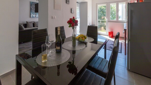 A modern dining area with a glass table, black chairs, a vase with red flowers, and a bottle of champagne. - Apartment IVA MAN (Crikvenica)