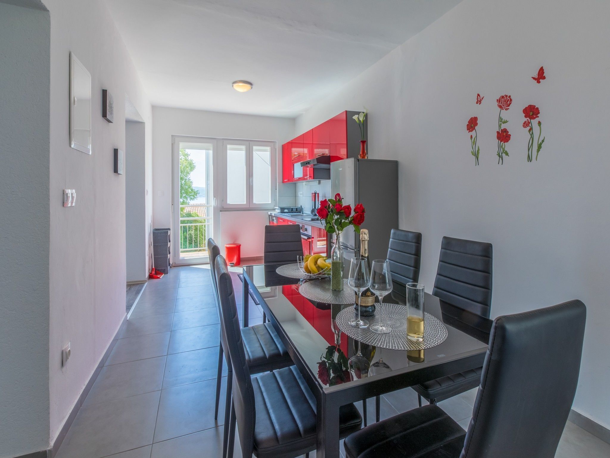A modern kitchen with red cabinets, a dining area with a glass table, and a balcony with a red chair.
