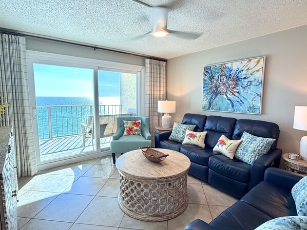 A spacious living room with a balcony, a blue sofa set, a round coffee table, and a large abstract painting on the wall.
