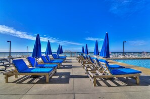 A beachside lounge area with rows of wooden lounge chairs and blue cushions, overlooking a clear blue sea.