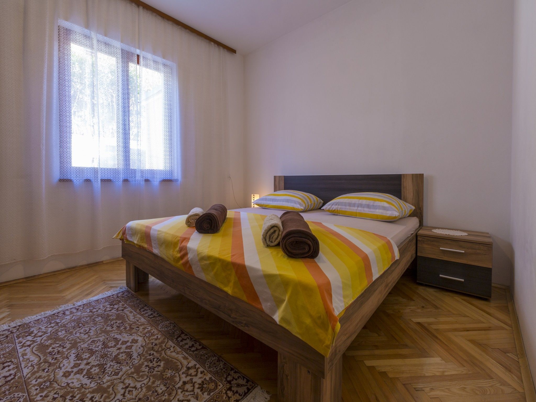 Une chambre à coucher avec un lit en bois, une literie à rayures, deux oreillers, une table de chevet et un tapis à motifs.