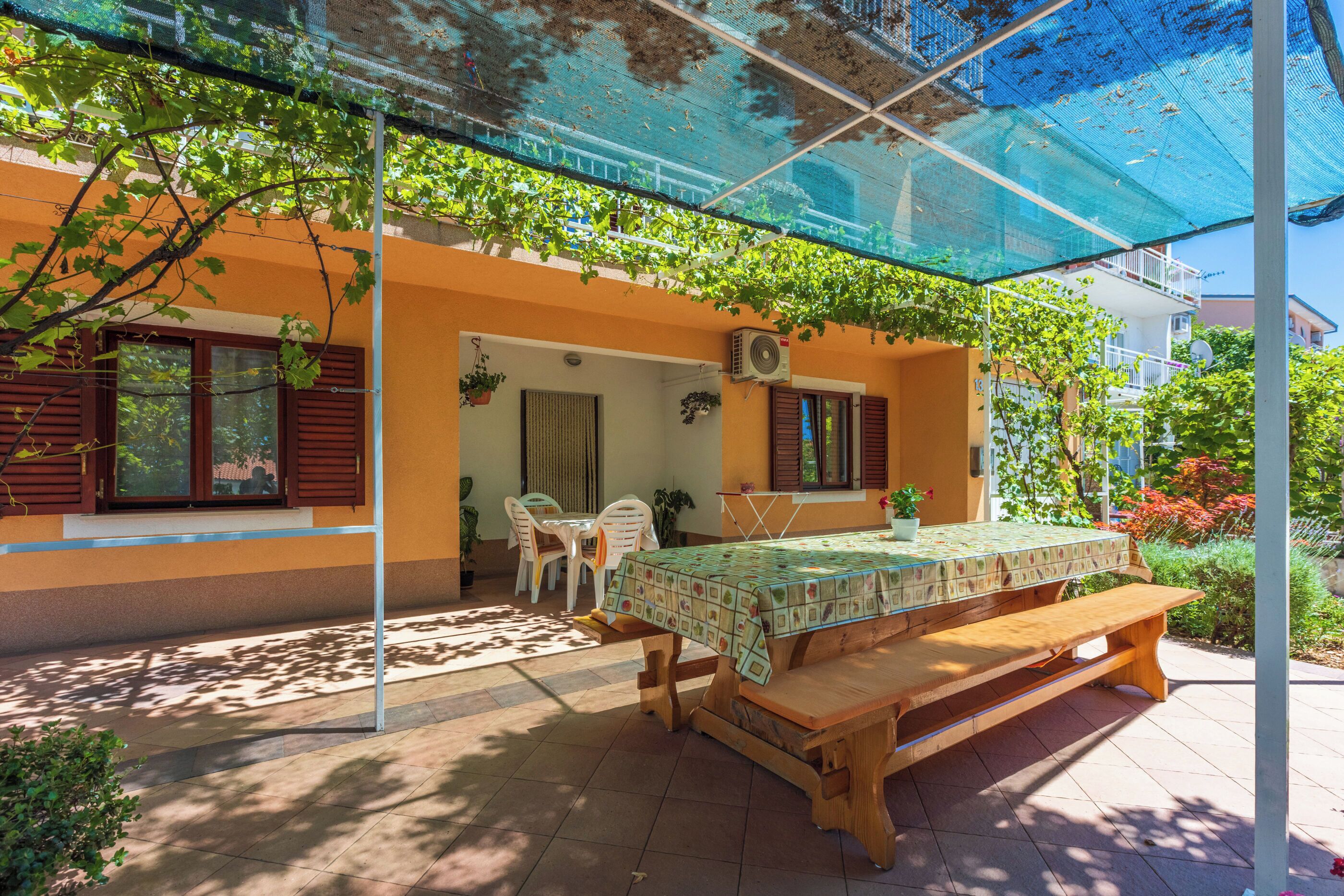 An outdoor patio with a wooden table and benches, covered by a blue canopy, surrounded by greenery and a building with orange walls.
