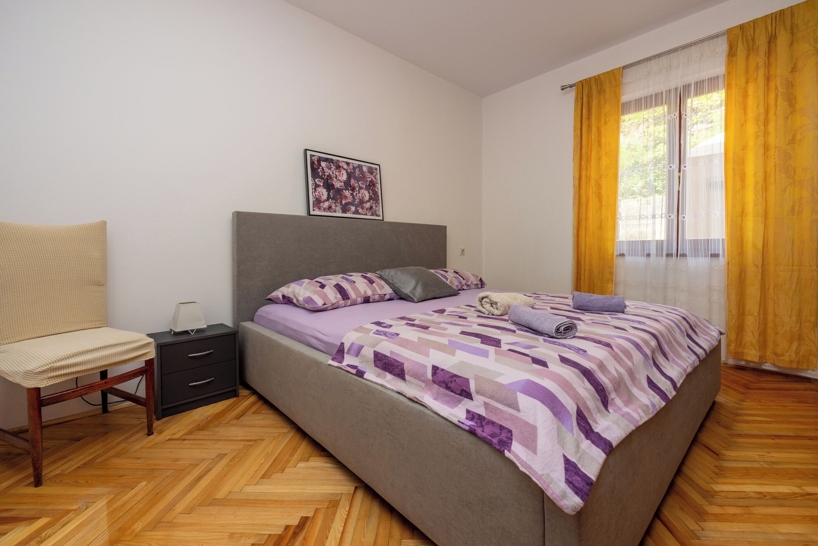 A bedroom with a bed, a nightstand, a chair, and a window with curtains.