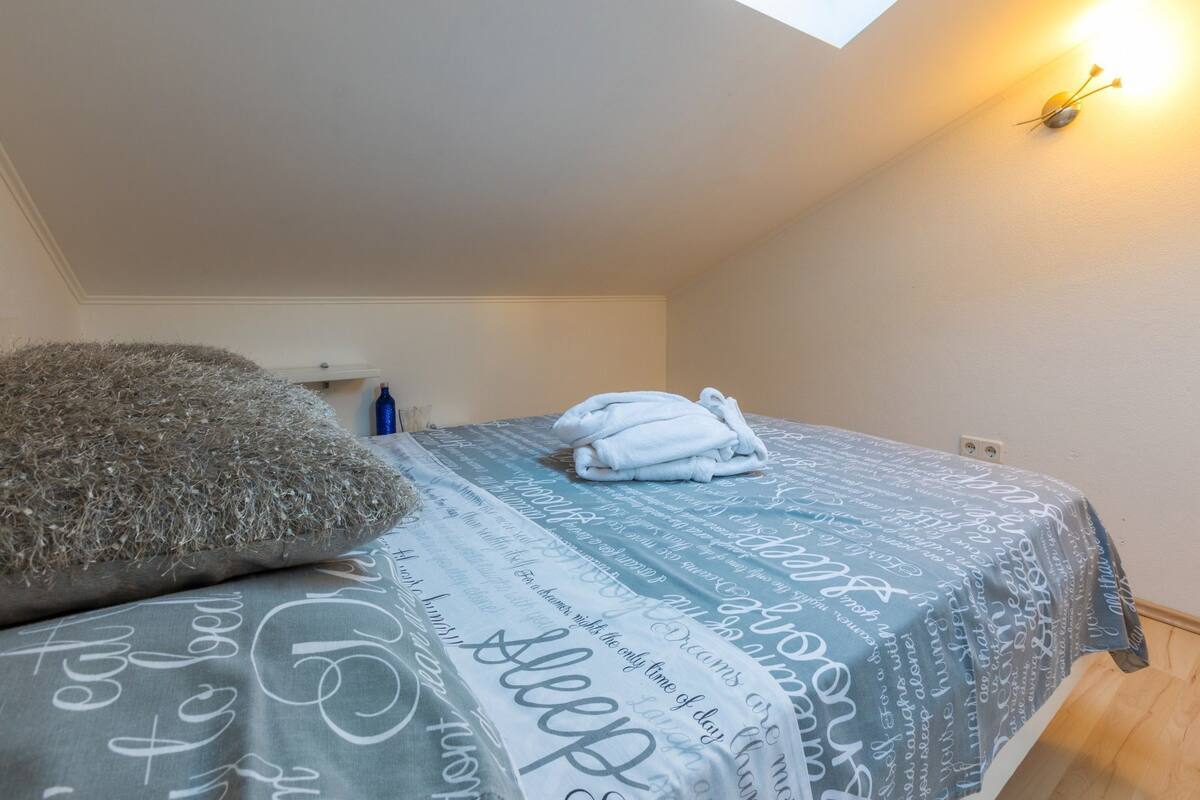 A bedroom with a skylight, a bed with a patterned bedspread, a pillow, and a bedside table with a bottle.