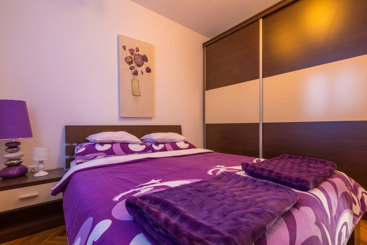 A bedroom with a large bed, a nightstand with a lamp, a wall-mounted artwork, and a sliding door closet.