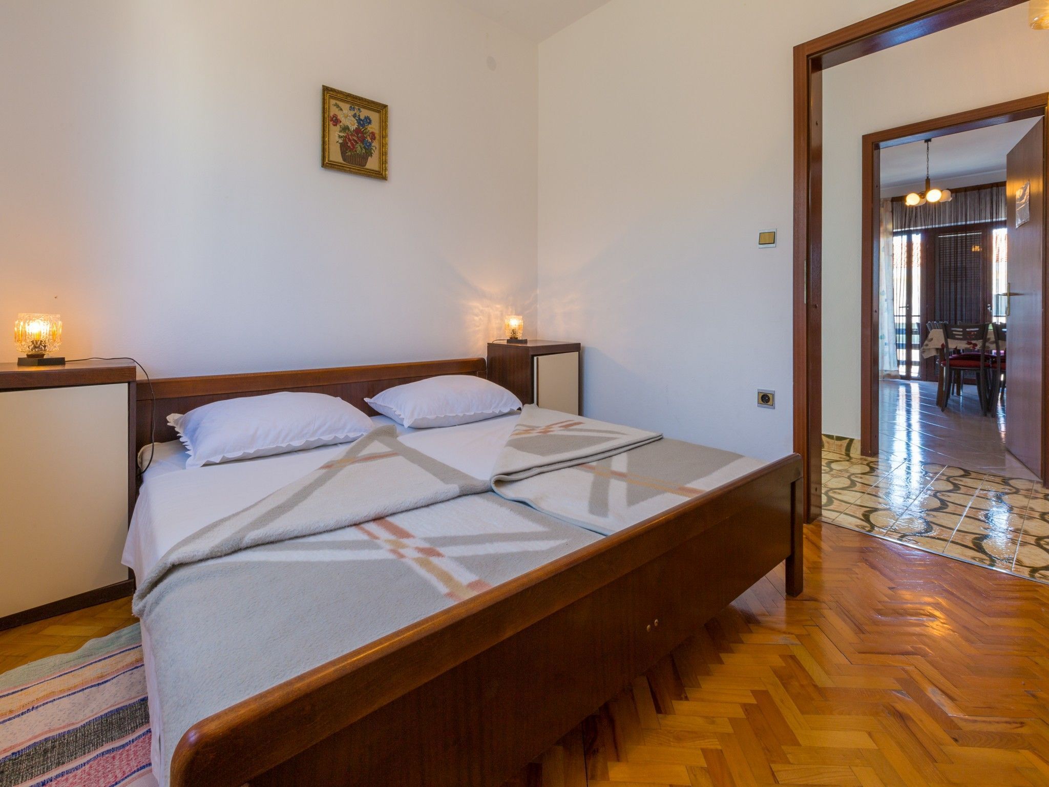Un lit bien fait, avec du linge de lit blanc et une couverture à motifs, une table de chevet avec une lampe, et un petit tableau encadré accroché au mur.