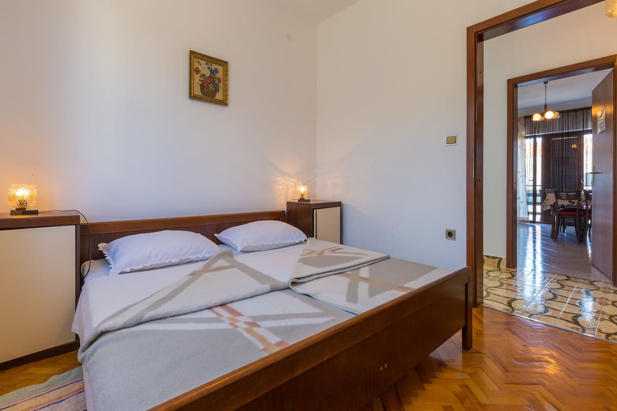 Una cama bien hecha con sábanas blancas y una manta estampada, una mesita de noche con una lámpara y un pequeño cuadro en la pared.