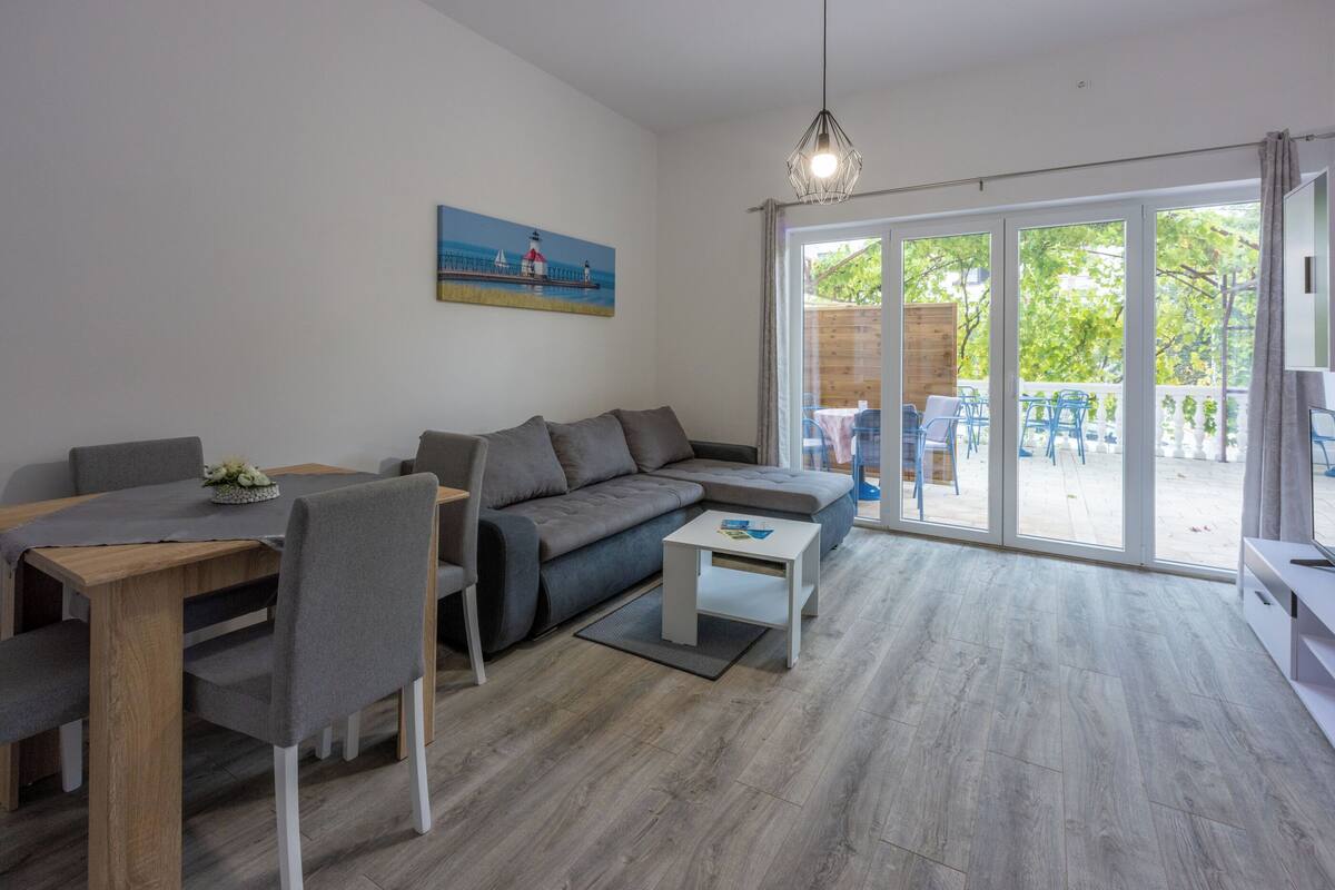 A modern living room with a sofa, dining table, and chairs, a painting on the wall, and large sliding glass doors leading to an outdoor area.