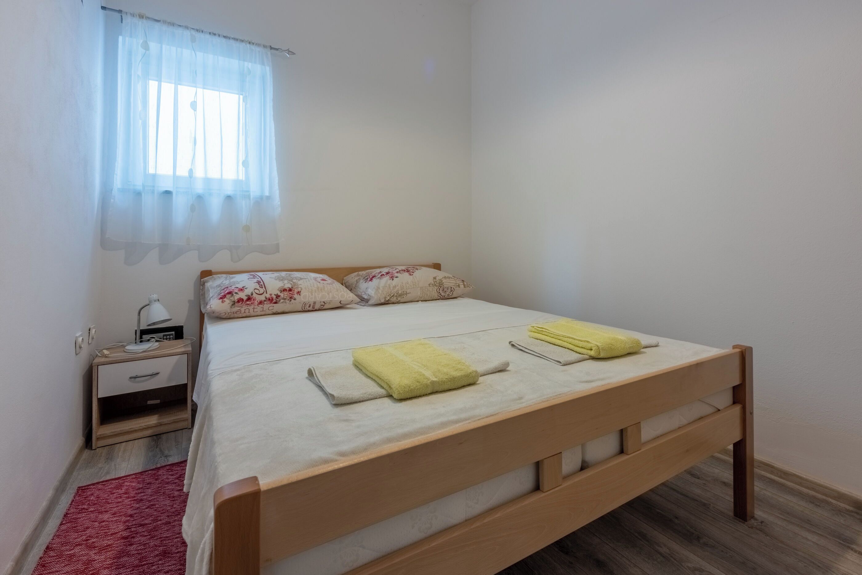 Uma cama bem arrumada com duas almofadas e duas toalhas dobradas, uma mesa de cabeceira com abajur, e uma janela com cortinas translúcidas.