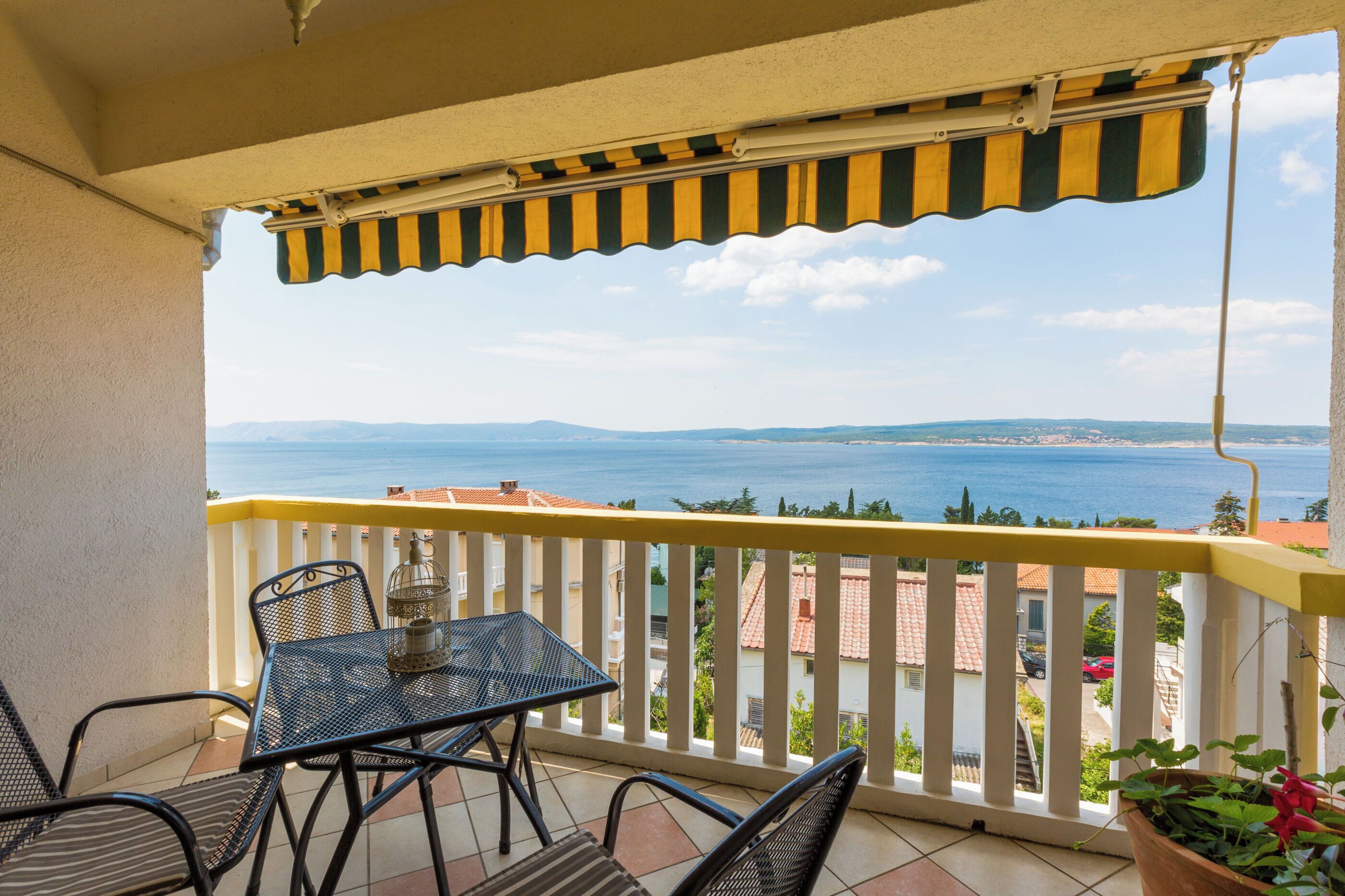 A balcony with a table and chairs, overlooking a coastal view with buildings and a clear sky.