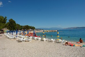 A pebble beach with rows of white lounge chairs and umbrellas, a lifeguard stand, and a clear view of the sea and distant mountains. - Apartments Neno (Crikvenica)