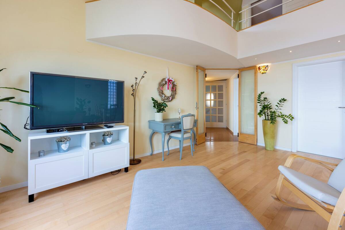A modern living room with a flat-screen TV, a sofa, a desk with a chair, and a large potted plant.