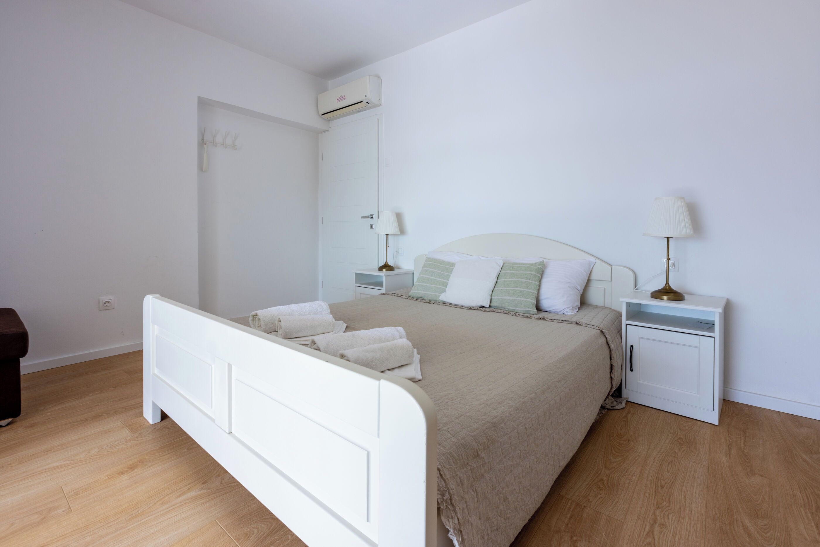 A bedroom with a white bed, a nightstand with a lamp, and an air conditioning unit on the wall.