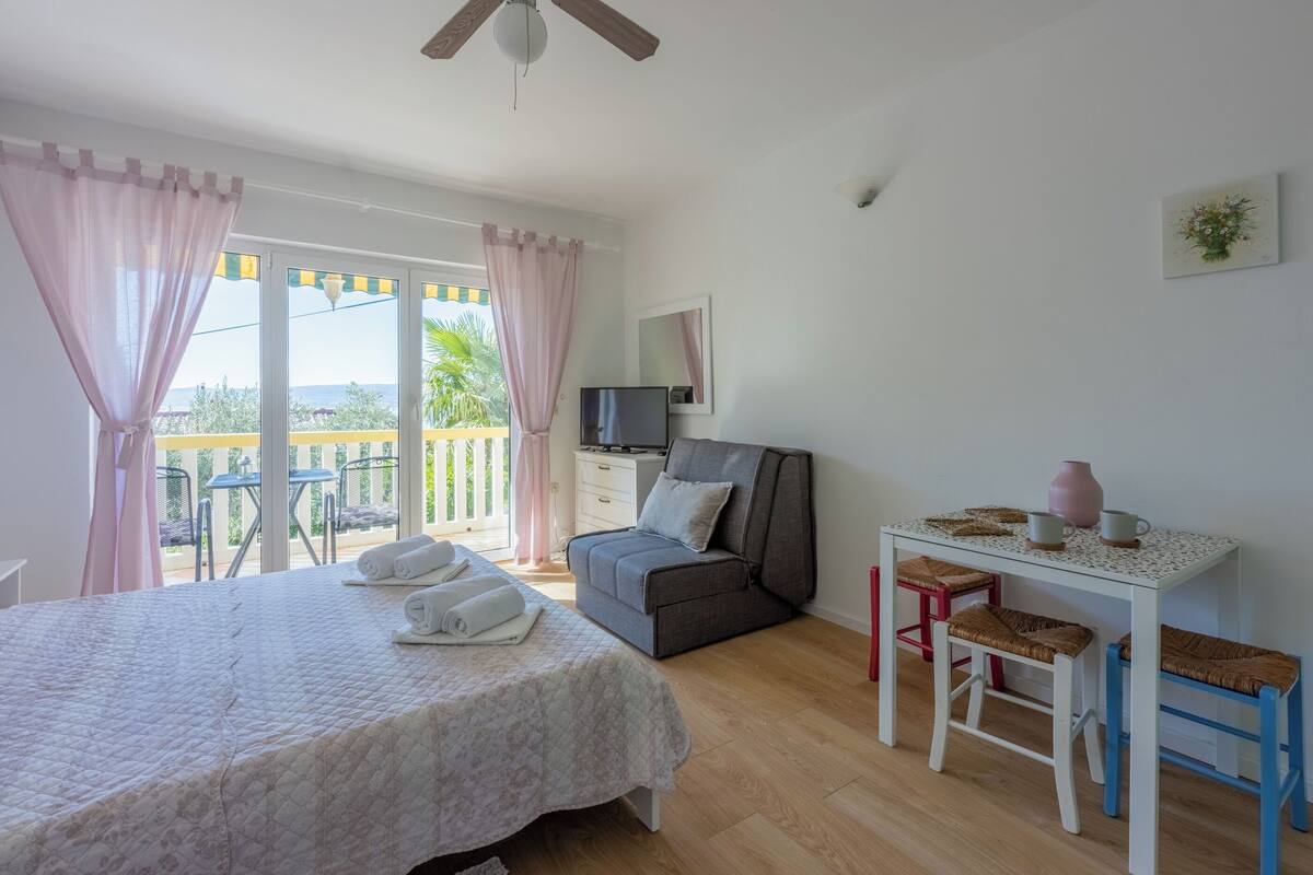 A bedroom with a bed, a desk, a chair, a TV, and a balcony with curtains.