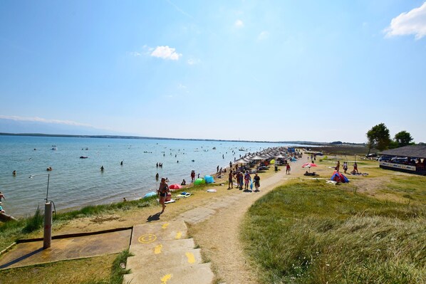 Ein Strand mit blauem Himmel, Menschen beim Schwimmen und Spazierengehen am Ufer, und ein Pfad, der zum Wasser führt.