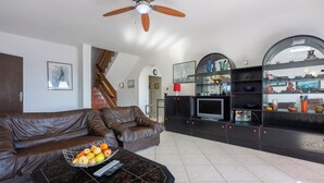 A modern living room with a brown leather sofa, a black coffee table with fruit and remote controls, a TV, and a decorative shelf with various items.