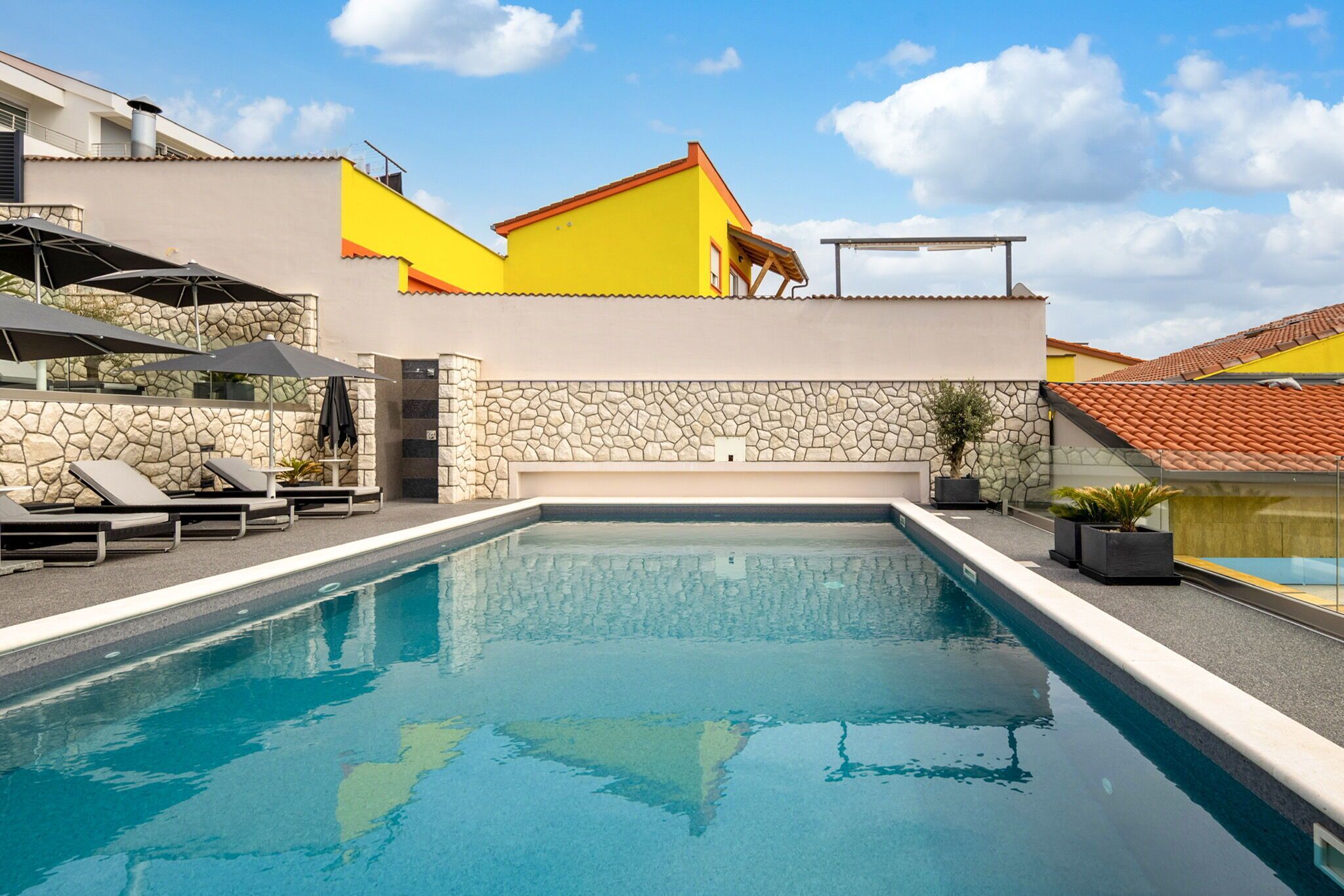 Une piscine avec des chaises longues et des parasols, un mur en pierre et un bâtiment jaune avec un toit rouge.
