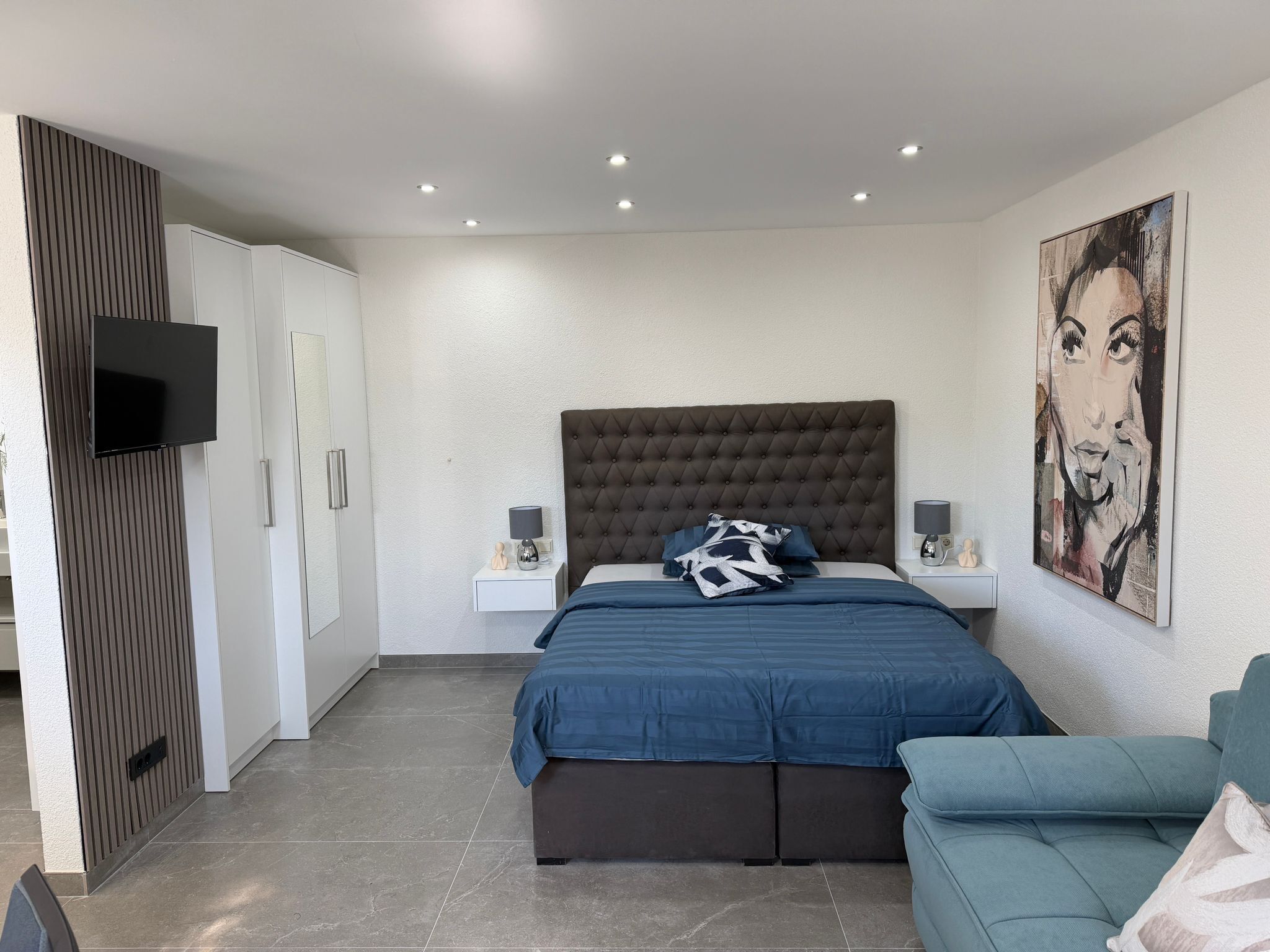 A modern bedroom with a large bed, a flat-screen TV, a tufted headboard, a painting on the wall, and a blue sofa.