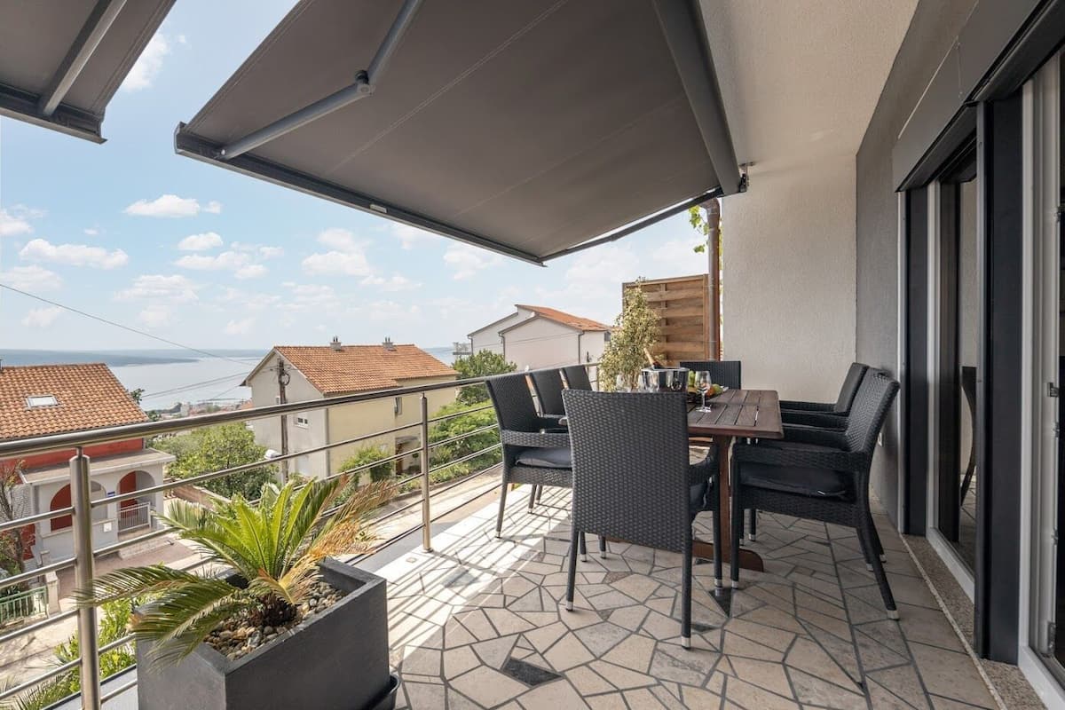 A balcony with a table and chairs, a potted plant, and a view of the sea and buildings.