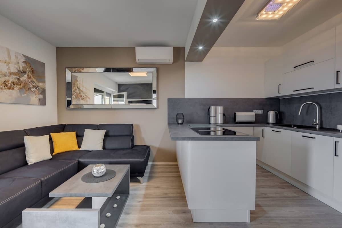 A modern apartment with a dark-colored sofa, a small coffee table, a kitchen with white cabinets and a black countertop, and a large mirror on the wall.