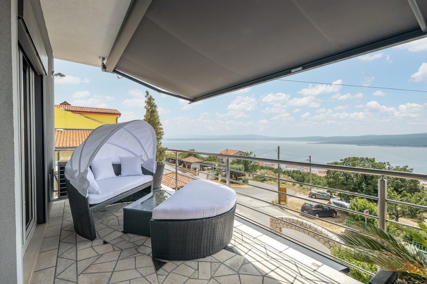 Een balkon met een rieten bank, een poef, een glazen tafel en uitzicht op zee en gebouwen.