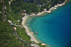 Une vue aérienne d’une zone côtière, avec une route qui serpente à travers une forêt et mène à une plage où l’on aperçoit des parasols et des personnes.