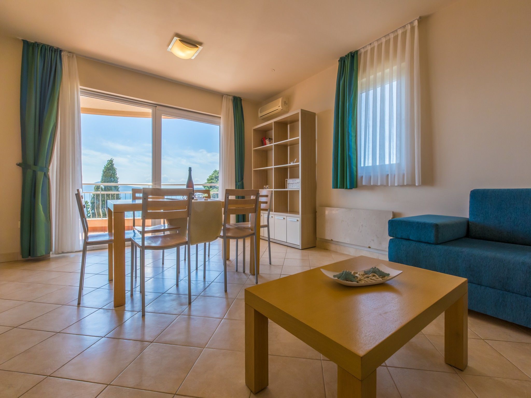 Ein Wohnzimmer mit Esstisch, Stühlen, einer Couch, einem Bücherregal und Blick ins Freie.